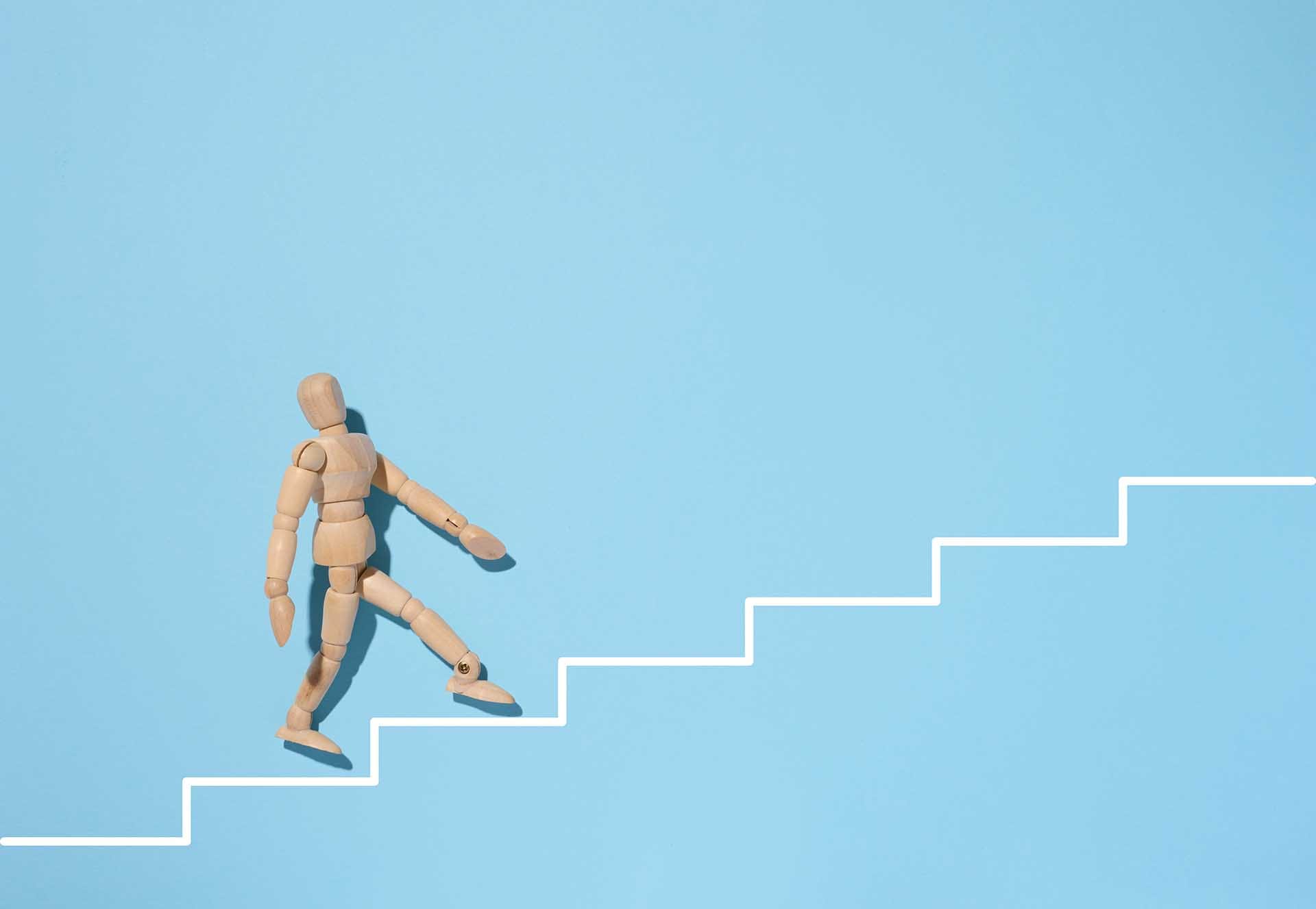 A wooden mannequin goes up a painted white staircase on a blue background. The concept of achieving the goal, overcoming obstacles